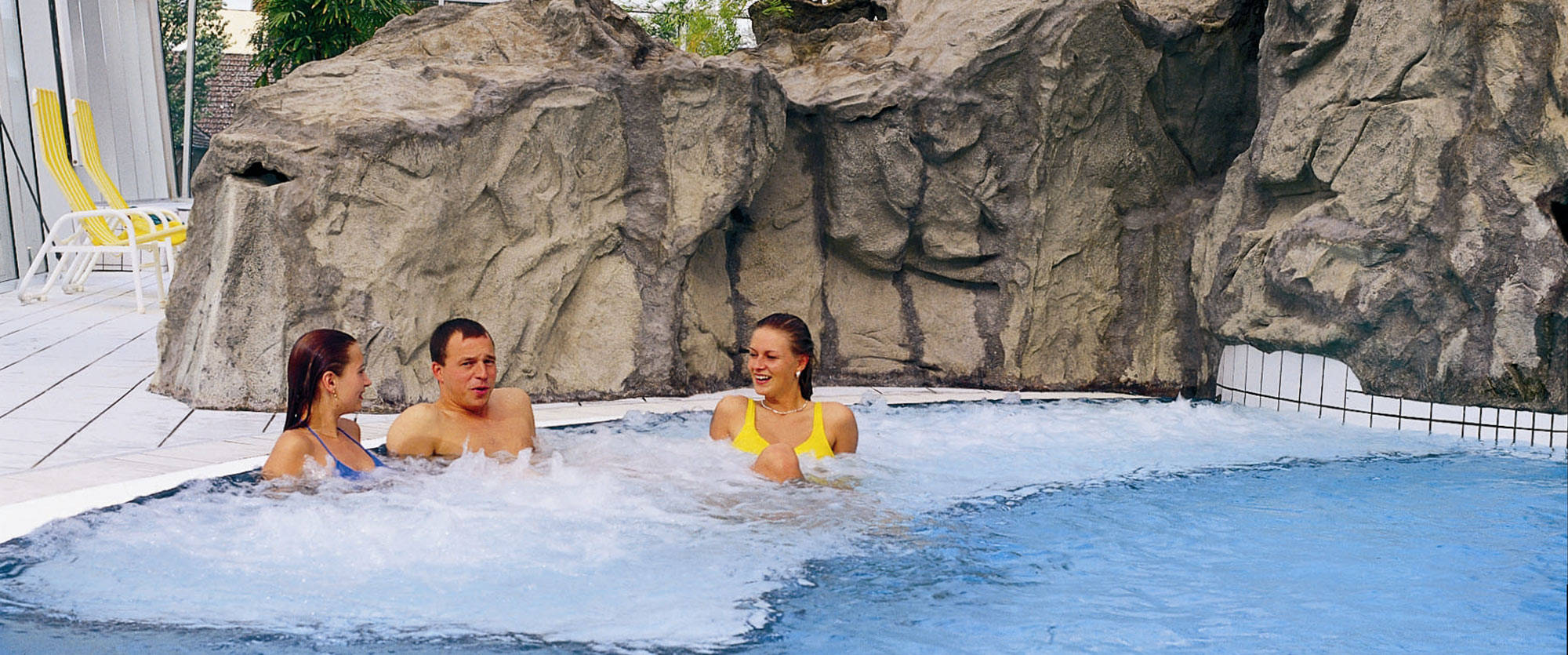 Swimming pool with swimmers on an air bubble recliner in front of artificial rocks